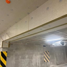 SAVRAN Sp.z o.o. - Ocieplenie stropu garażu wełną mineralną, widoczne płyty przymocowane do sufitu za pomocą okrągłych łączników, instalacje elektryczne pod sufitem, betonowe ściany z pasami ostrzegawczymi.