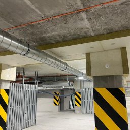SAVRAN Sp.z o.o. - Wnętrze parkingu podziemnego z widoczną instalacją wentylacyjną pod stropem, zabezpieczoną izolacją termiczną, oraz słupami oznaczonymi żółto-czarnymi pasami.