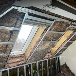 SAVRAN Sp.z o.o. - Ocieplenie poddasza wełną mineralną, widoczne metalowe profile konstrukcyjne i okno dachowe z widokiem na drzewa.