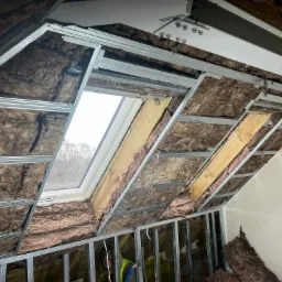 Ocieplenie poddasza wełną mineralną, widoczne metalowe profile konstrukcyjne i okno dachowe z widokiem na drzewa.