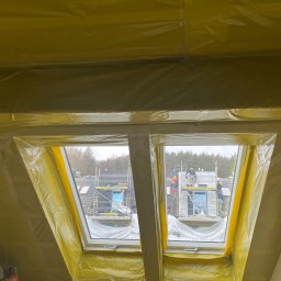 SAVRAN Sp.z o.o. - Poddasze zabezpieczone folią ochronną podczas prac budowlanych, widok z okna na rusztowania i pracowników na zewnątrz budynku.