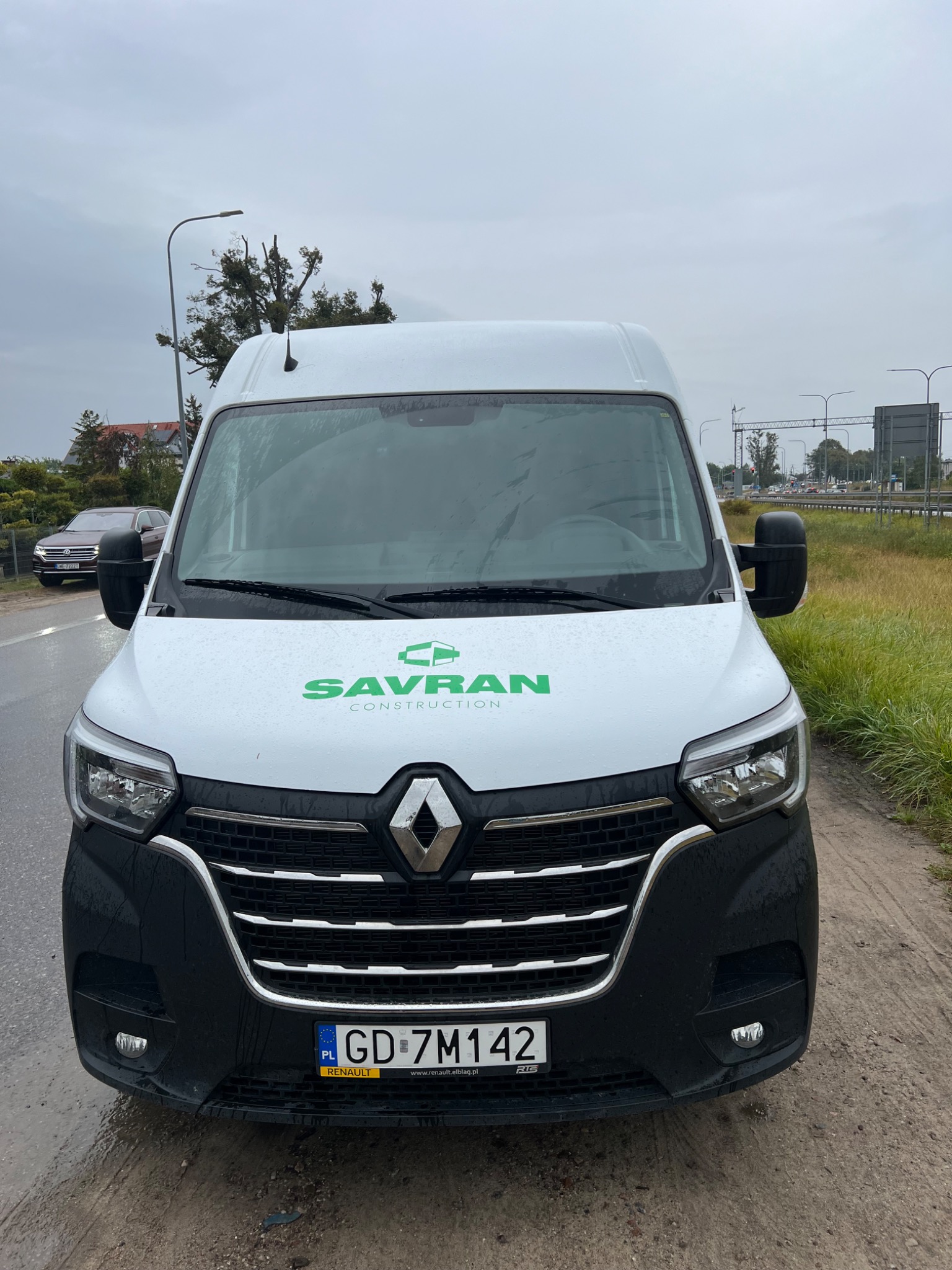Biały samochód dostawczy Renault Master z zielonym logo Savran Construction na masce, zaparkowany na poboczu drogi w pochmurny dzień.