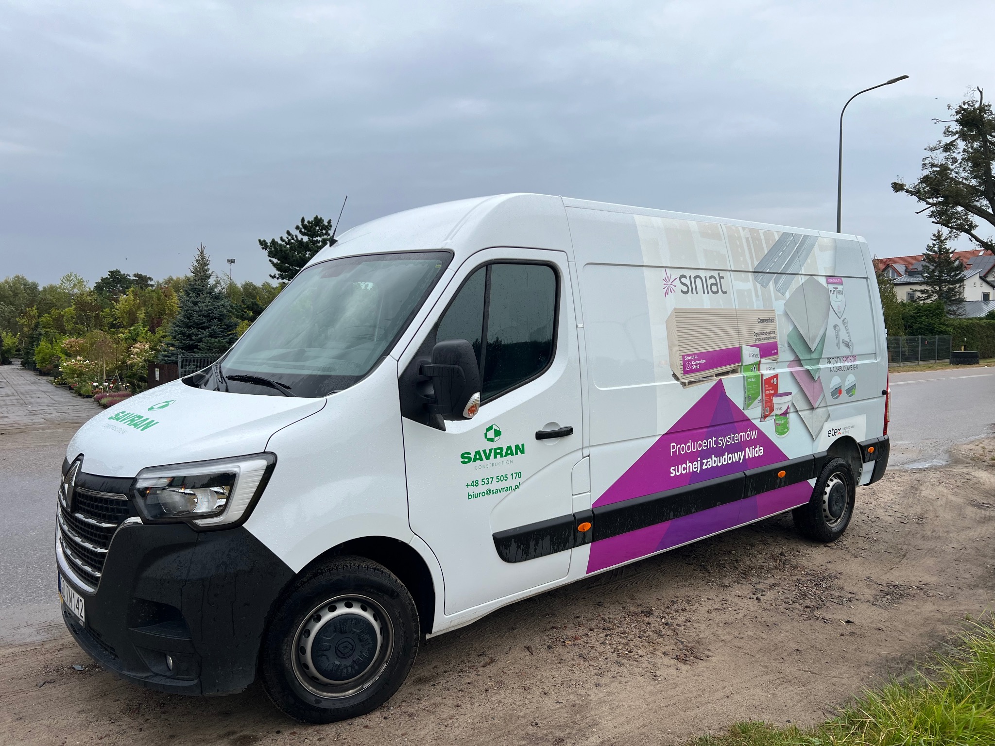 Biały samochód dostawczy firmy Savran Construction z reklamą systemów suchej zabudowy Nida, zaparkowany na poboczu w pochmurny dzień.