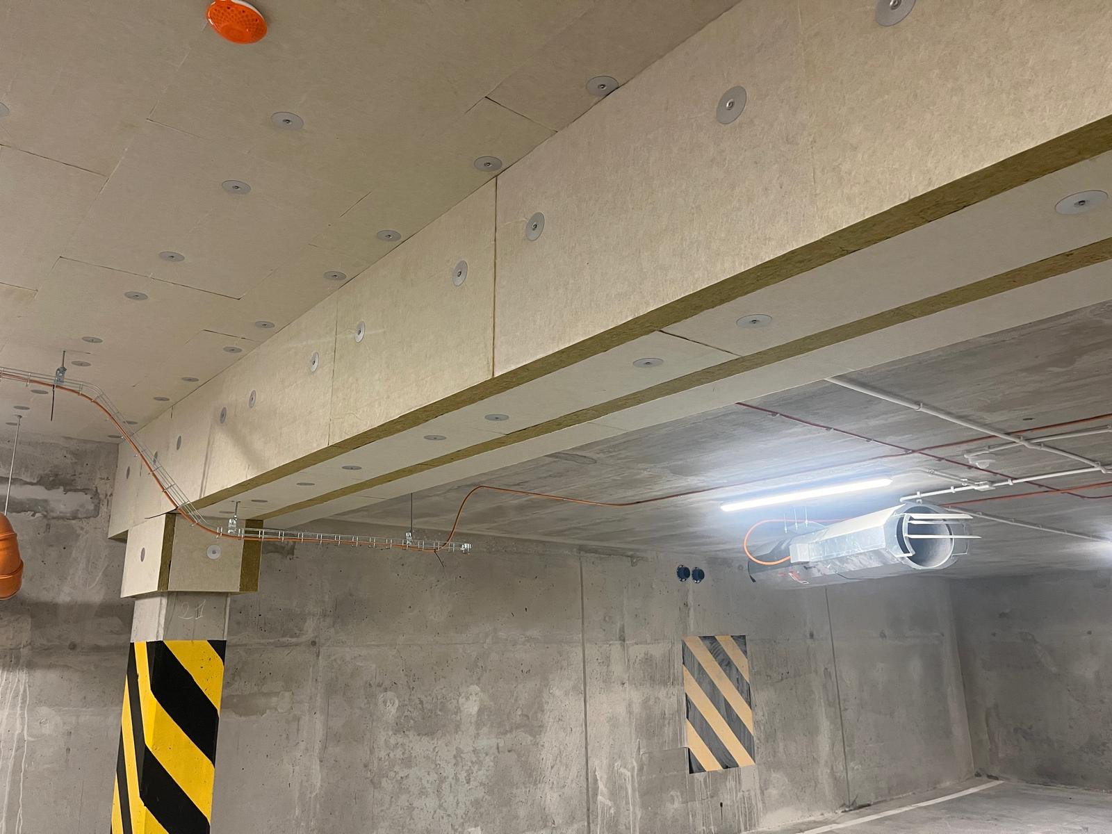 Ocieplenie stropu garażu wełną mineralną, widoczne płyty przymocowane do sufitu za pomocą okrągłych łączników, instalacje elektryczne pod sufitem, betonowe ściany z pasami ostrzegawczymi.