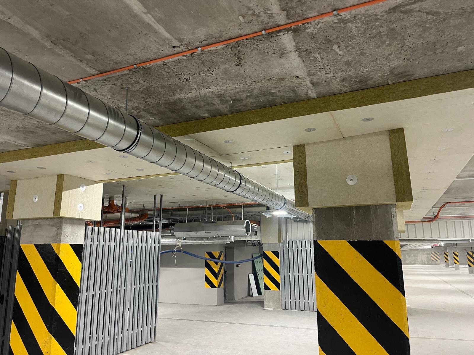Wnętrze parkingu podziemnego z widoczną instalacją wentylacyjną pod stropem, zabezpieczoną izolacją termiczną, oraz słupami oznaczonymi żółto-czarnymi pasami.