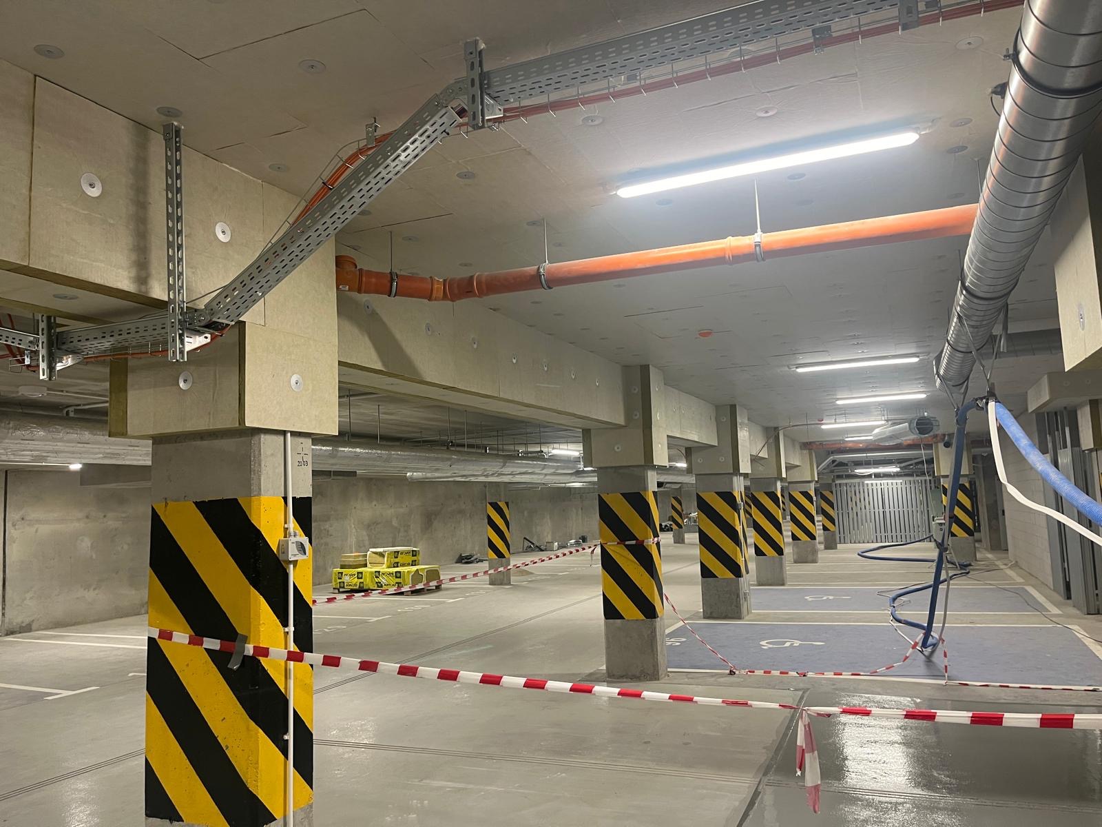 Wnętrze parkingu podziemnego z widocznymi elementami instalacji wentylacyjnych i elektrycznych pod sufitem, zabezpieczone słupy w pasy ostrzegawcze, oraz materiały budowlane na palecie w tle.