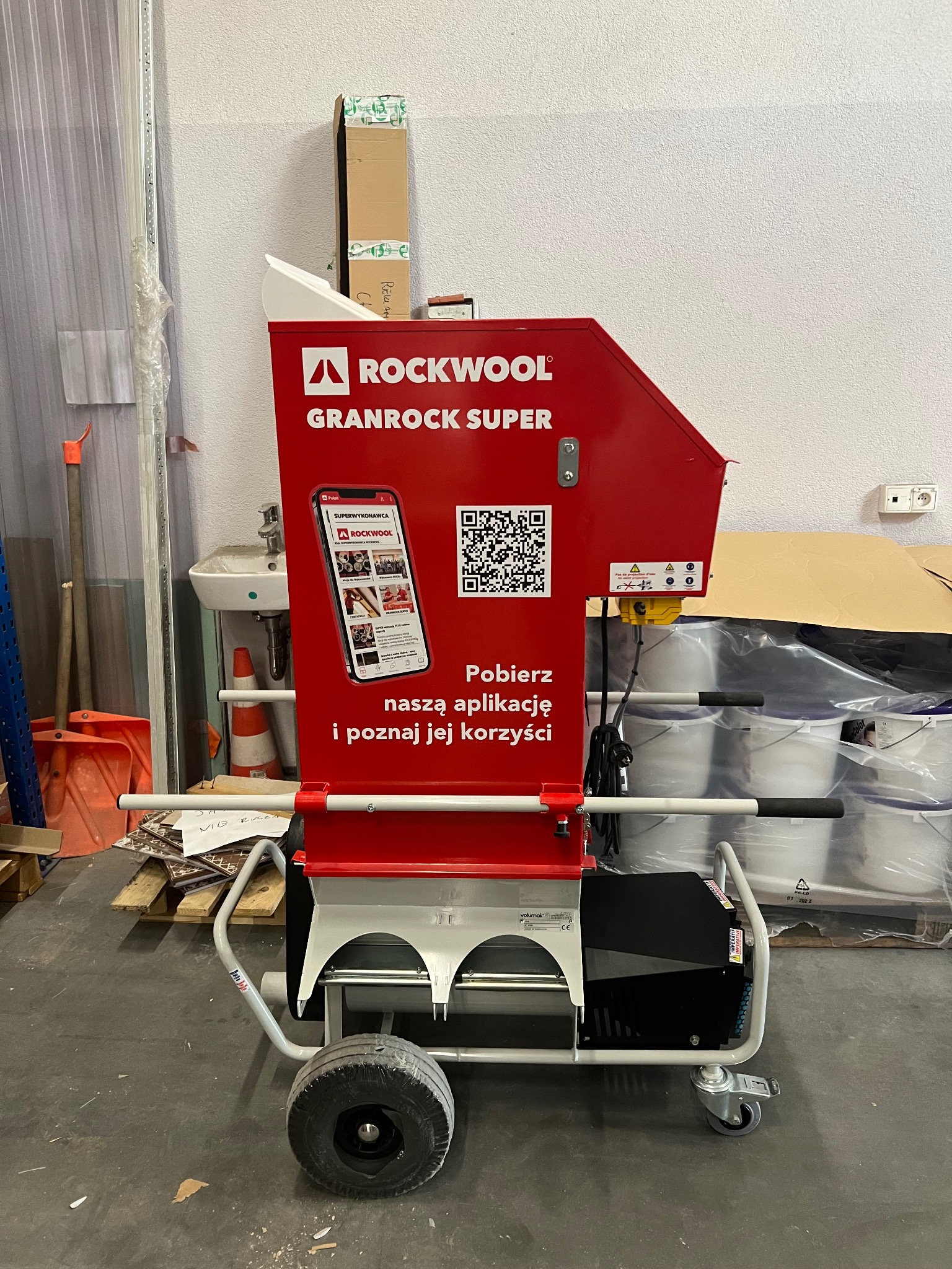 Czerwona maszyna do granulatu Rockwool Granrock Super z naklejką reklamującą aplikację mobilną, na tle magazynu z wiadrami i materiałami budowlanymi.
