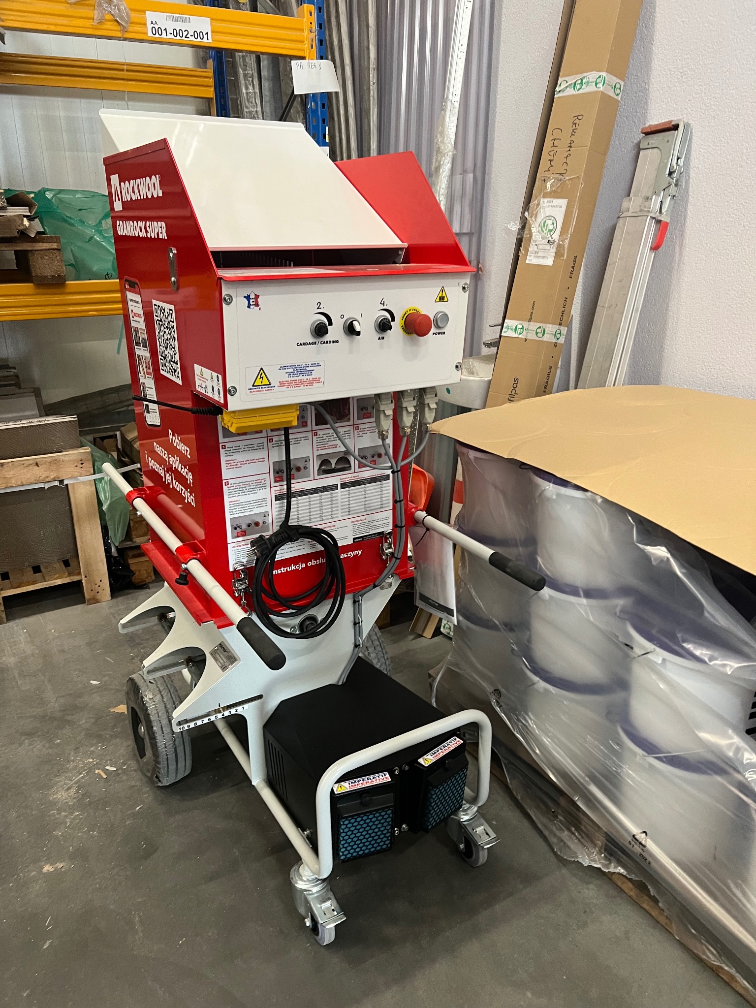 Maszyna do granulowania wełny mineralnej Rockwool Granrock Super, widoczna w magazynie z paletami i opakowaniami materiałów budowlanych, z instrukcjami obsługi i ostrzeżeniami na obudowie.