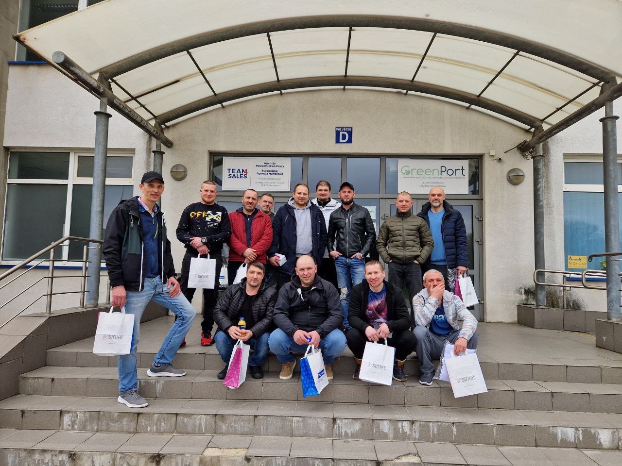 Grupowe zdjęcie pracowników budowlanych przed budynkiem biurowym GreenPort, trzymających torby z logo Siniat.