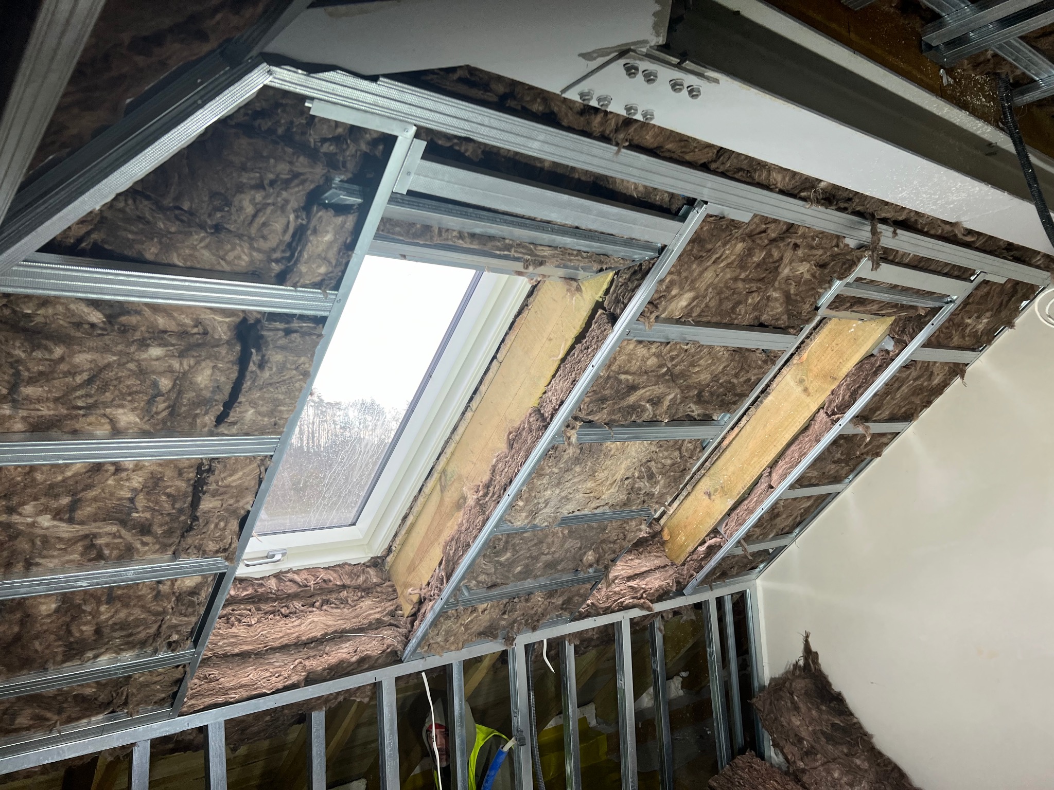 Ocieplenie poddasza wełną mineralną, widoczne metalowe profile konstrukcyjne i okno dachowe z widokiem na drzewa.