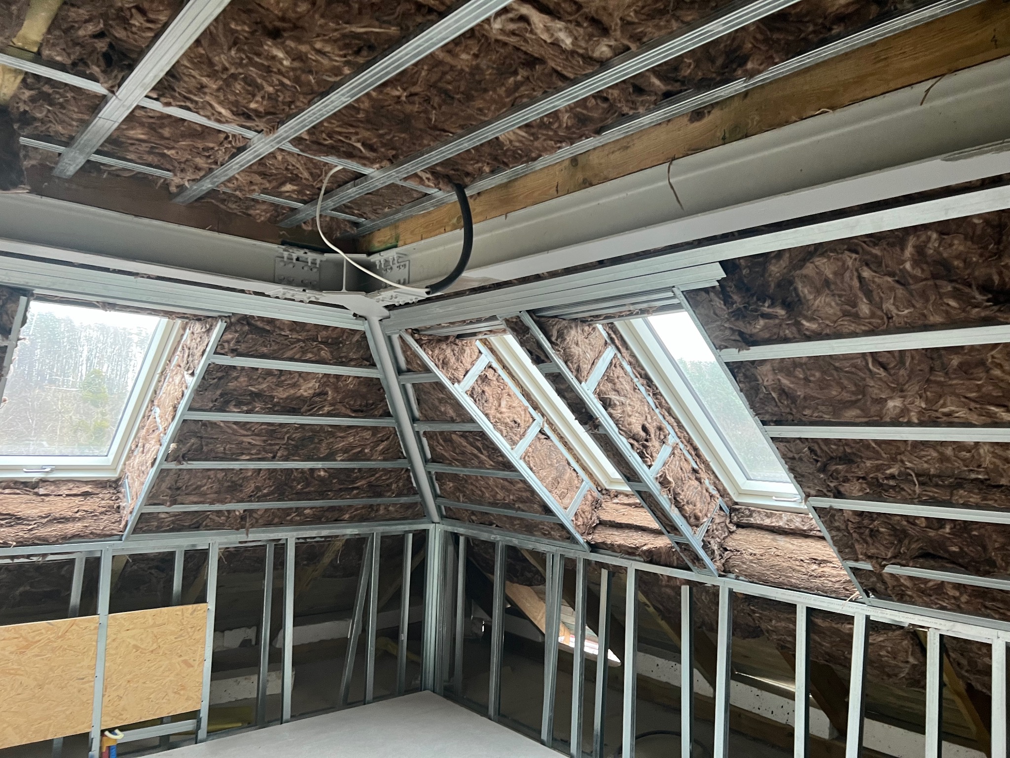 Skośne poddasze z oknami dachowymi w trakcie izolacji wełną mineralną i montażu metalowej konstrukcji pod płyty gipsowo-kartonowe, widoczne kable elektryczne.