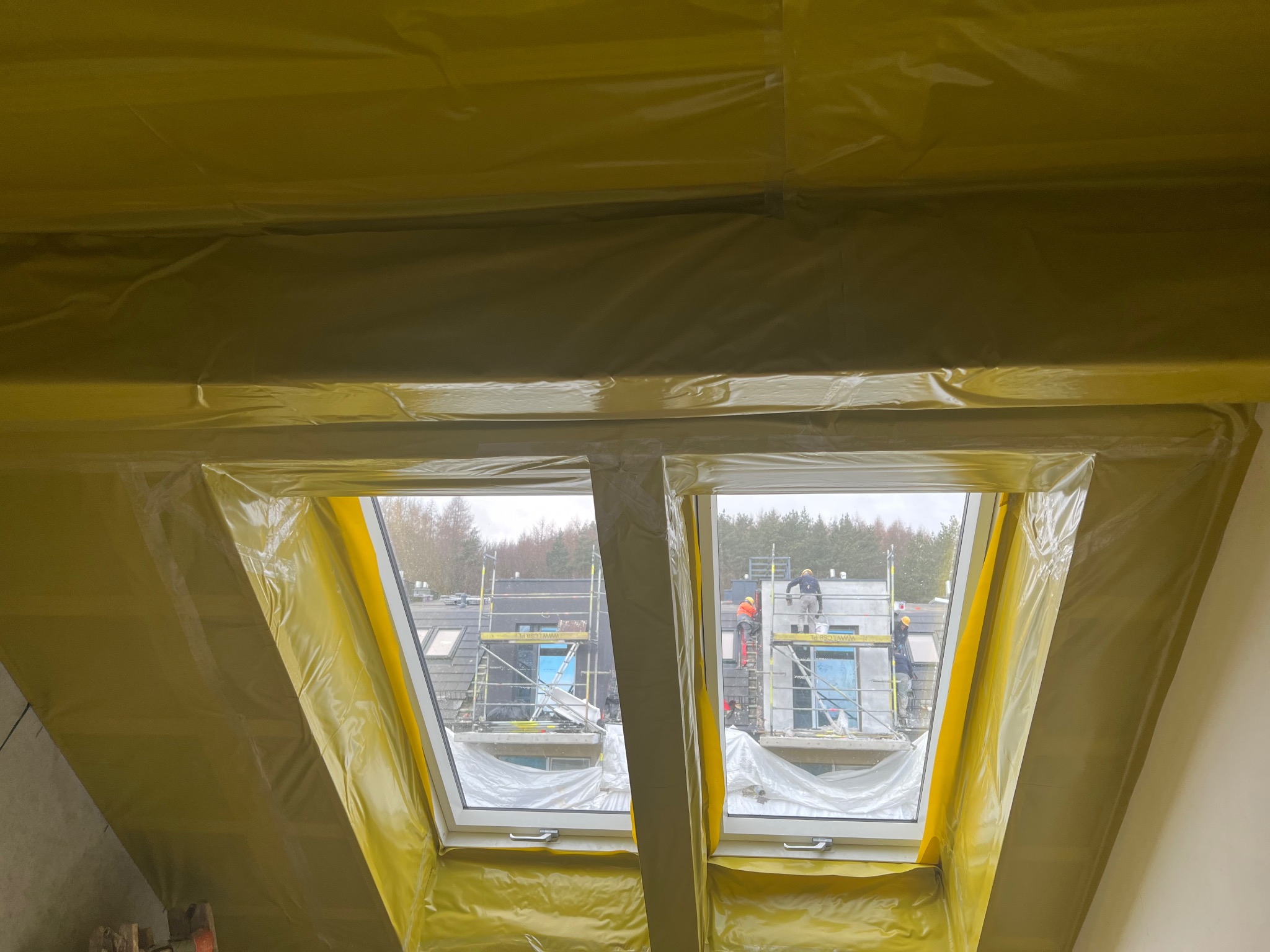 Poddasze zabezpieczone folią ochronną podczas prac budowlanych, widok z okna na rusztowania i pracowników na zewnątrz budynku.