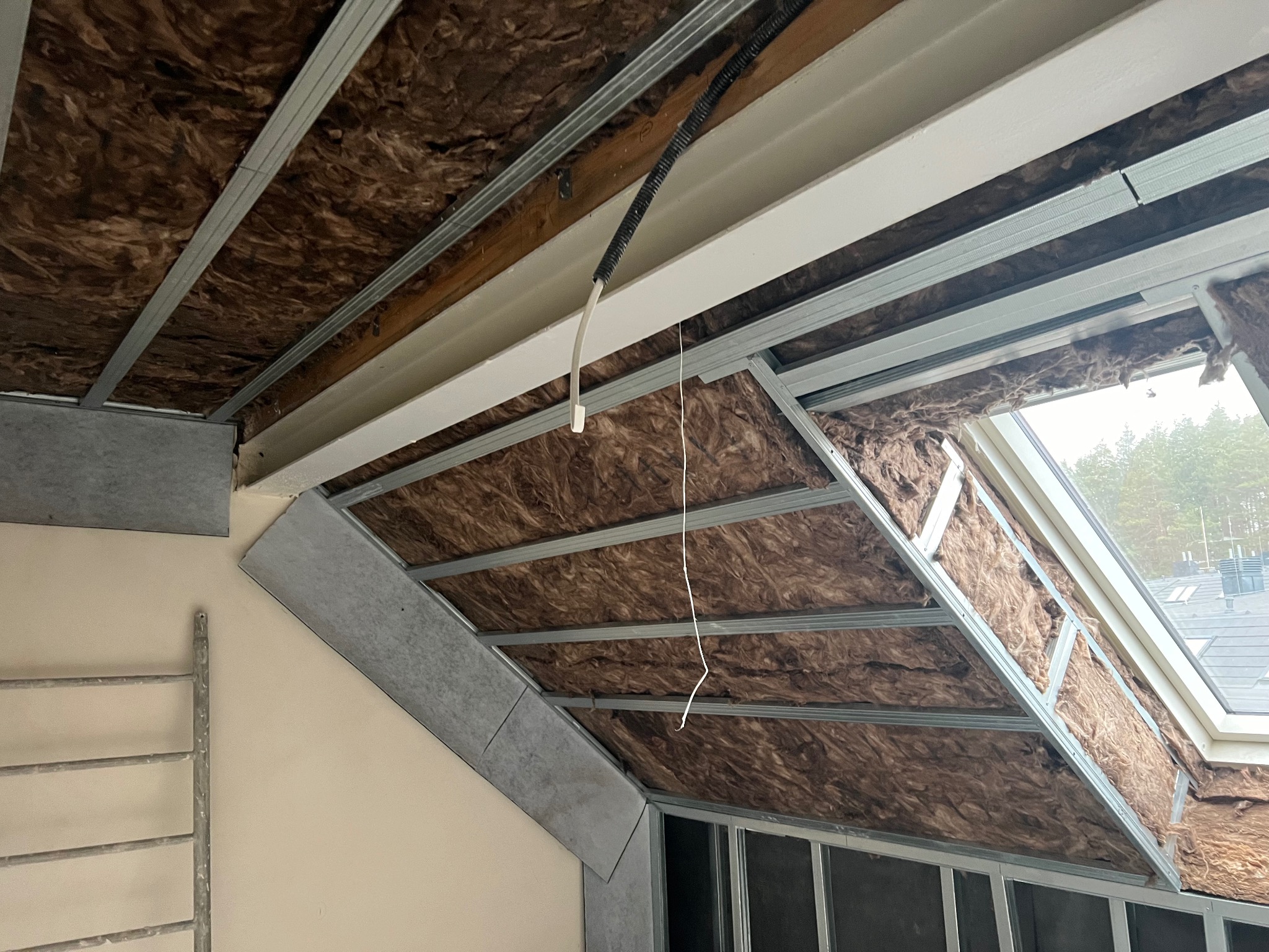 Skośne poddasze w trakcie ocieplania wełną mineralną, widoczne metalowe profile konstrukcyjne i częściowo zamontowane płyty kartonowo-gipsowe przy ścianie, drabina oparta o ścianę w lewym dolnym rogu.