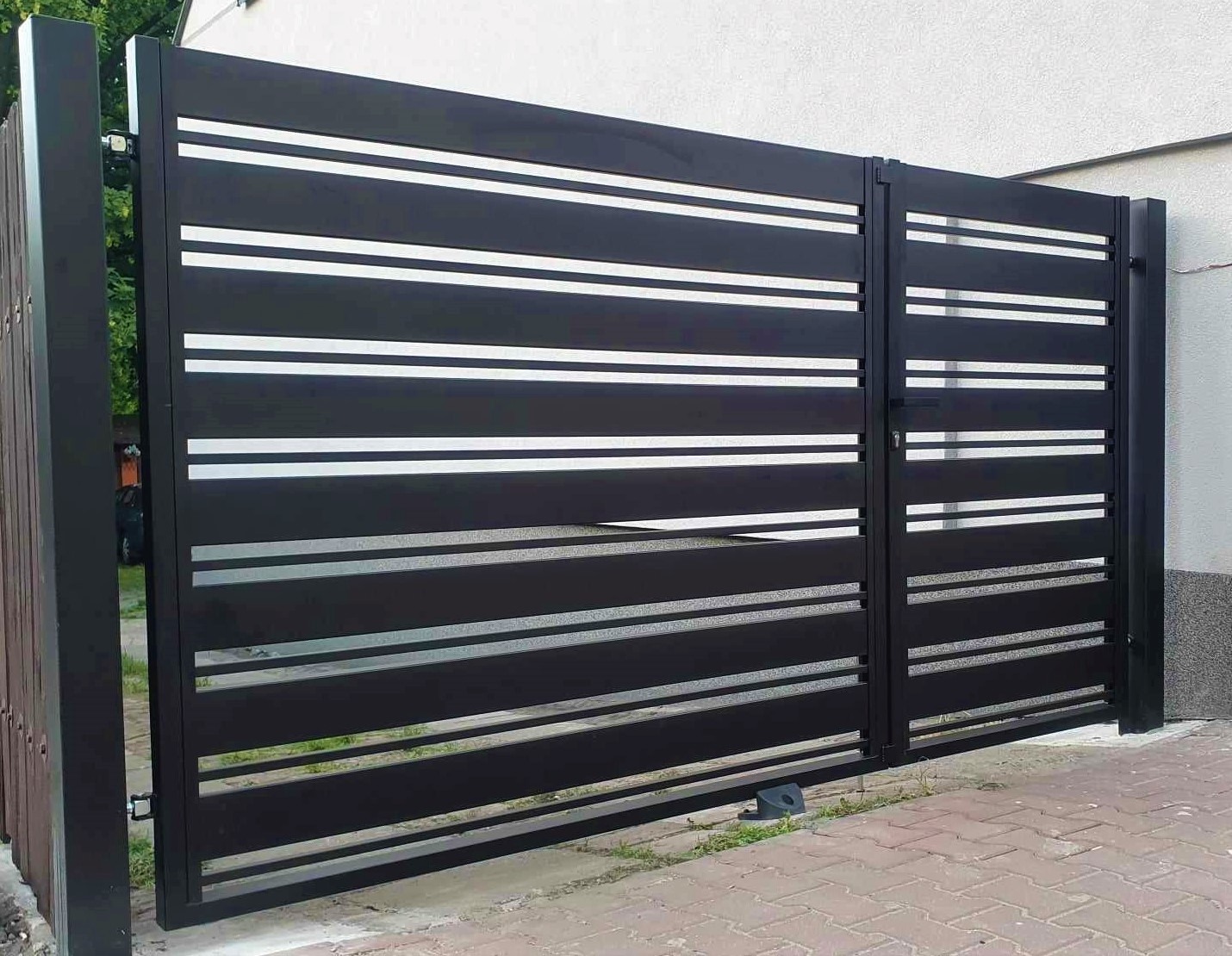 Nowoczesna, czarna brama wjazdowa z poziomymi, srebrnymi akcentami, zamontowana na posesji z kostką brukową i jasnym budynkiem w tle.