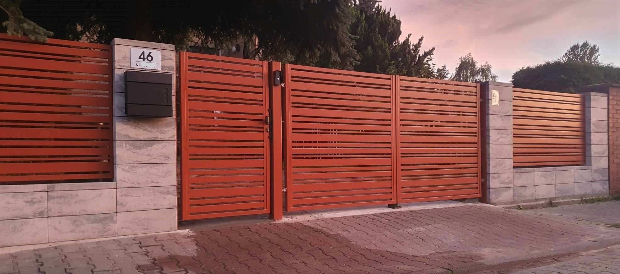 Nowoczesna, pozioma brama wjazdowa z metalowymi lamelami w kolorze ceglastym, zamontowana na słupkach z szarej kostki elewacyjnej, z czarną skrzynką na listy i numerem domu na słupku.