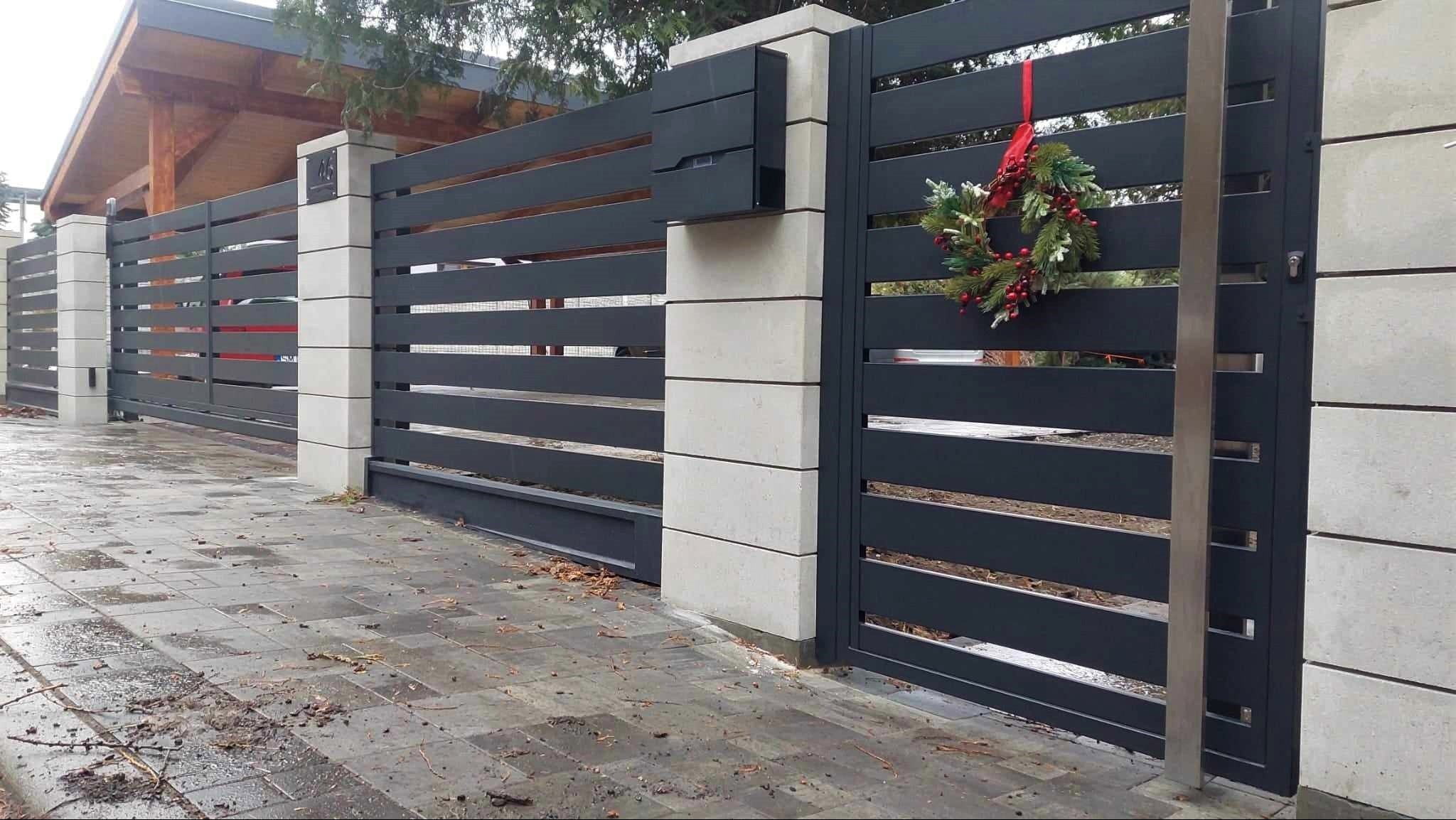 Nowoczesna, przesuwna brama wjazdowa z szarych, poziomych paneli metalowych, osadzona na słupkach z jasnego betonu, z skrzynką na listy i świątecznym wieńcem.