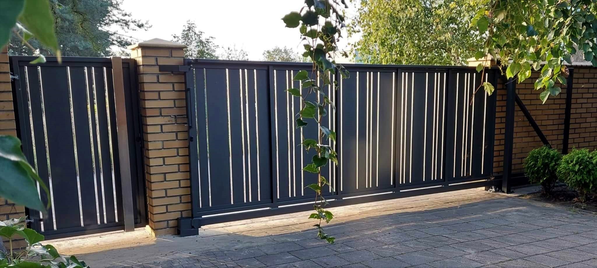 Nowoczesna, przesuwna brama wjazdowa z pionowymi, jasnymi szczeblinami, osadzona między ceglanymi słupkami i murem, z rośliną pnącą się po jej fragmencie.