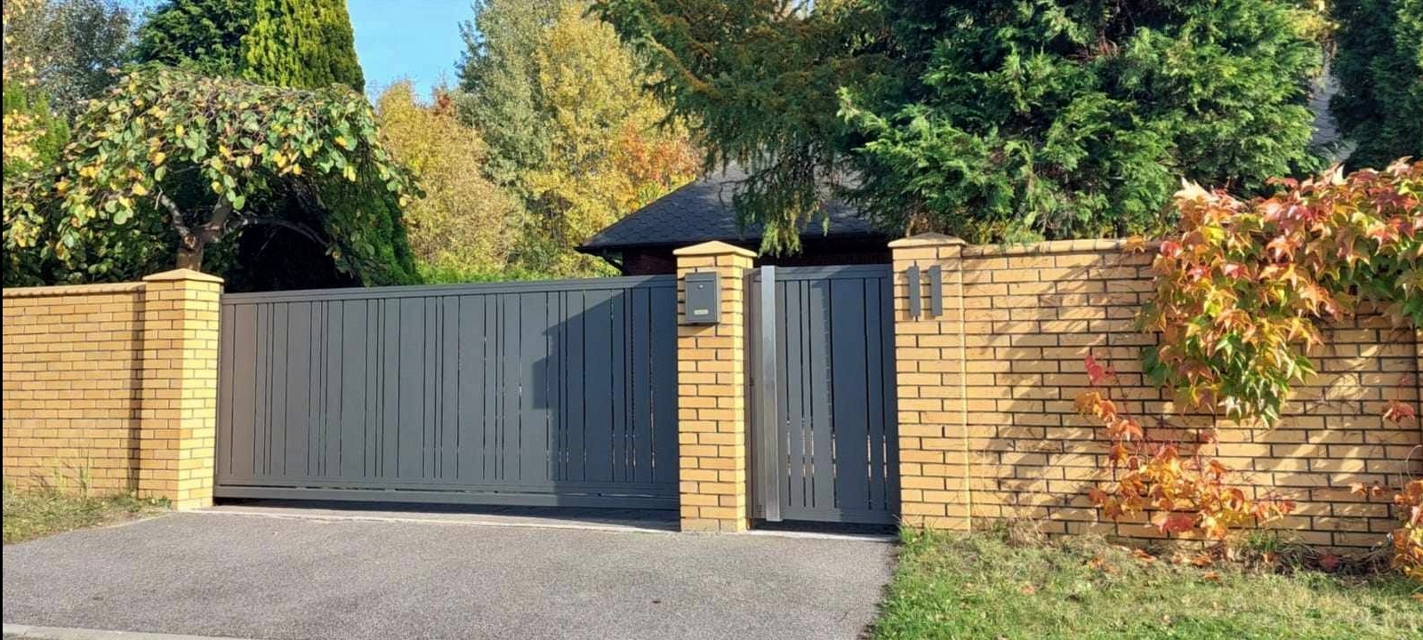 Przesuwna brama wjazdowa w kolorze antracytowym z furtką, osadzona w murze z żółtej cegły z skrzynką na listy, na tle jesiennego ogrodu.