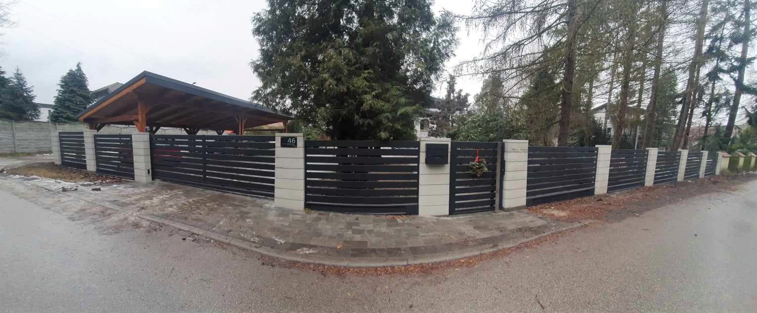 Nowoczesne ogrodzenie posesji z poziomych, czarnych paneli między jasnymi, betonowymi słupkami; na jednym ze słupków skrzynka pocztowa i świąteczny wianek.