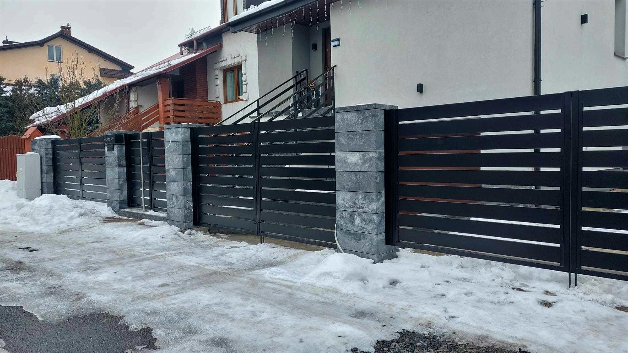 Nowoczesna brama wjazdowa i ogrodzenie panelowe w kolorze antracytowym z elementami drewnopodobnymi, osadzone na słupkach z szarego betonu architektonicznego, widok z zewnątrz posesji w zimowej...