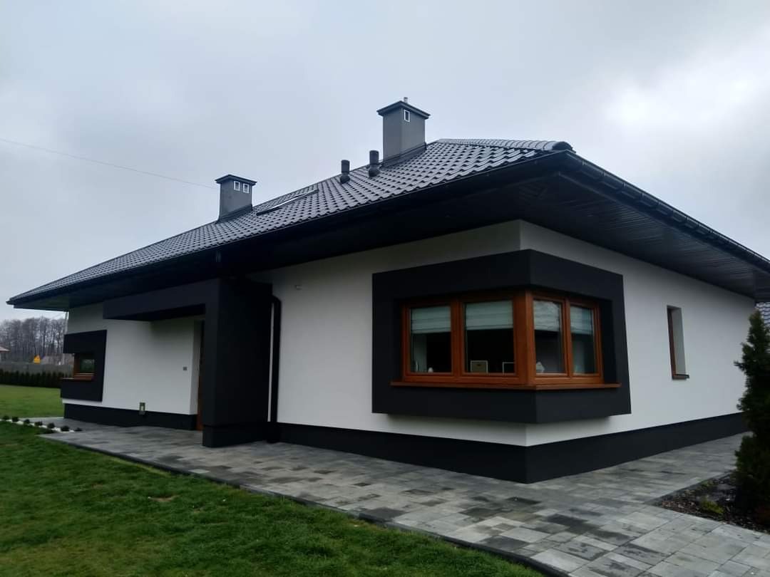 Nowoczesny dom jednorodzinny z białą elewacją i czarnymi akcentami, ciemna dachówka, widok z zewnątrz na szarym brukowanym chodniku i trawniku, pochmurne niebo.