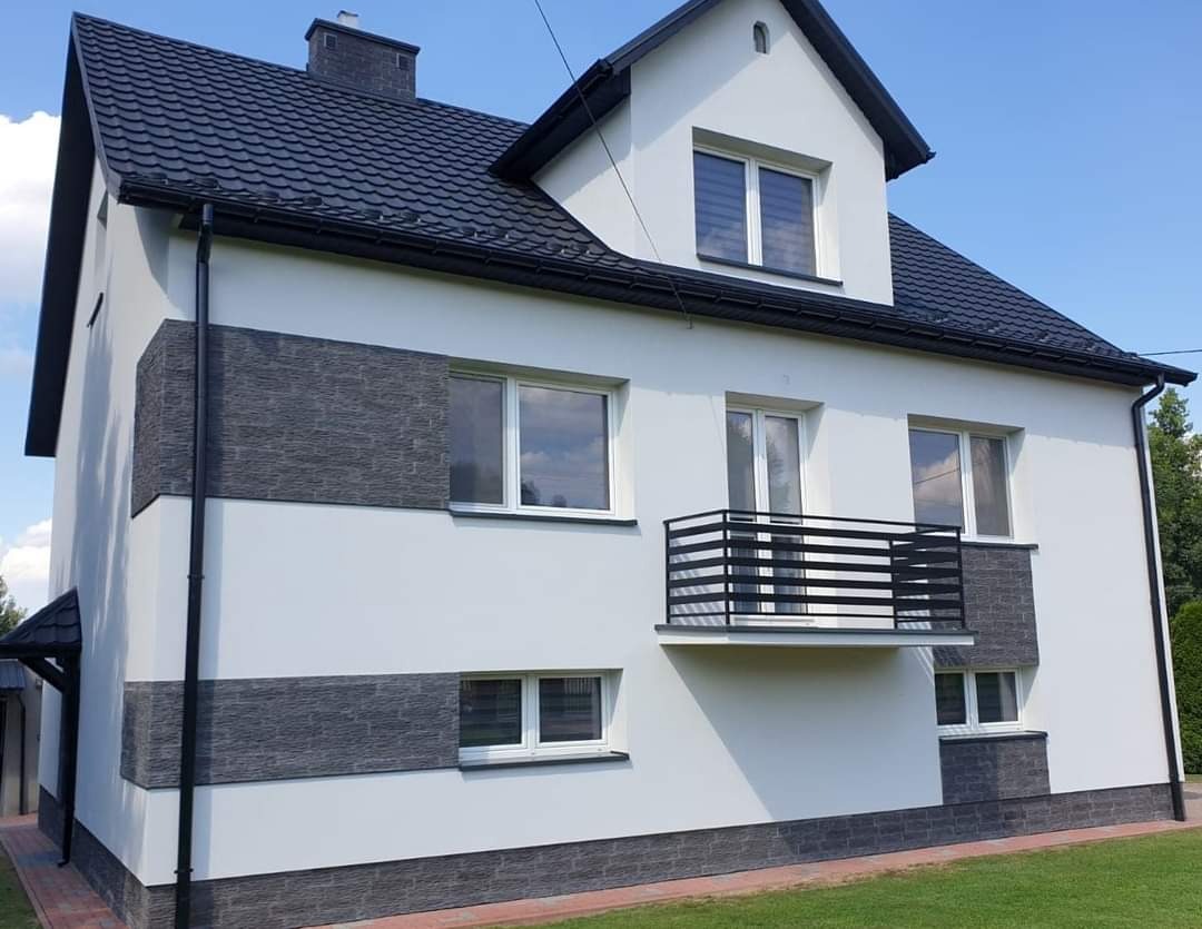 Dwukondygnacyjny dom z białą elewacją i ciemnoszarą dachówką, z balkonem z czarną balustradą i fragmentami elewacji wykończonymi dekoracyjnym kamieniem w odcieniach szarości.