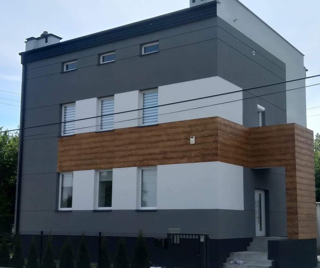 Dwukondygnacyjny dom po renowacji elewacji, z szarą i białą farbą oraz drewnopodobnym panelem na środkowej części, widoczny z niskiej perspektywy.