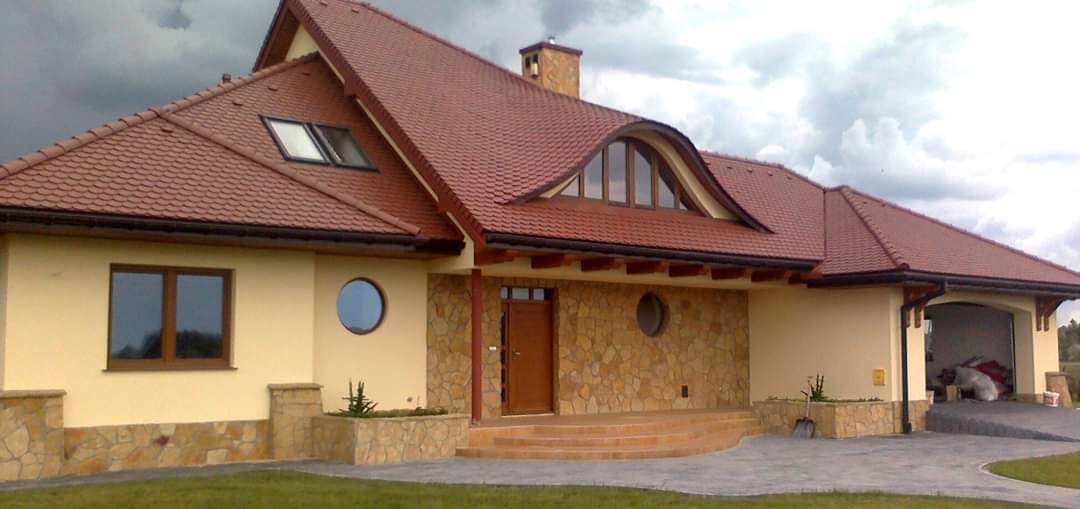 Nowoczesny dom jednorodzinny z beżową elewacją, kamiennym wykończeniem, czerwoną dachówką i okrągłymi oknami, widok na front budynku w pochmurny dzień.
