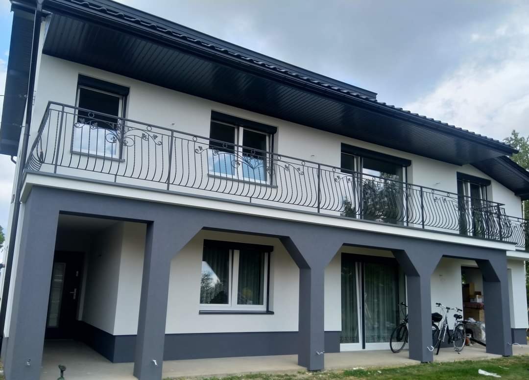 Dwukondygnacyjny dom z jasną elewacją i ciemnoszarą podmurówką, balkon z ozdobną, metalową balustradą, widoczne dwa rowery zaparkowane przed budynkiem.
