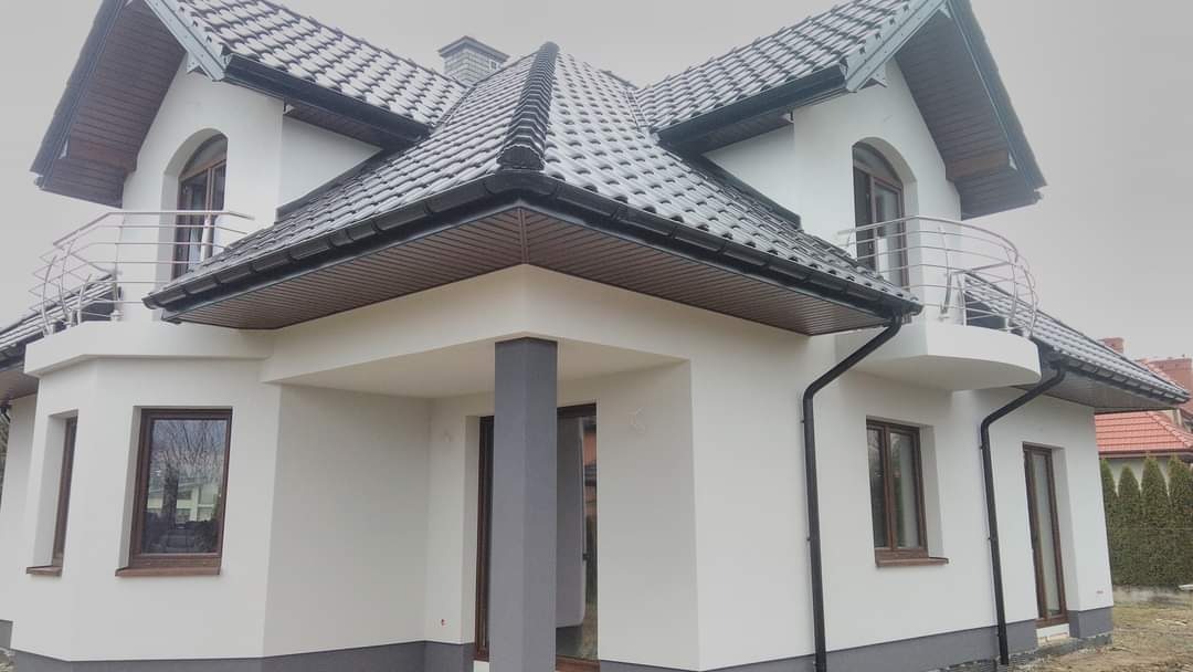 Nowoczesny dom jednorodzinny z białą elewacją, szarą podmurówką i grafitowym dachem z balkonami z metalową balustradą, widok z zewnątrz w pochmurny dzień.