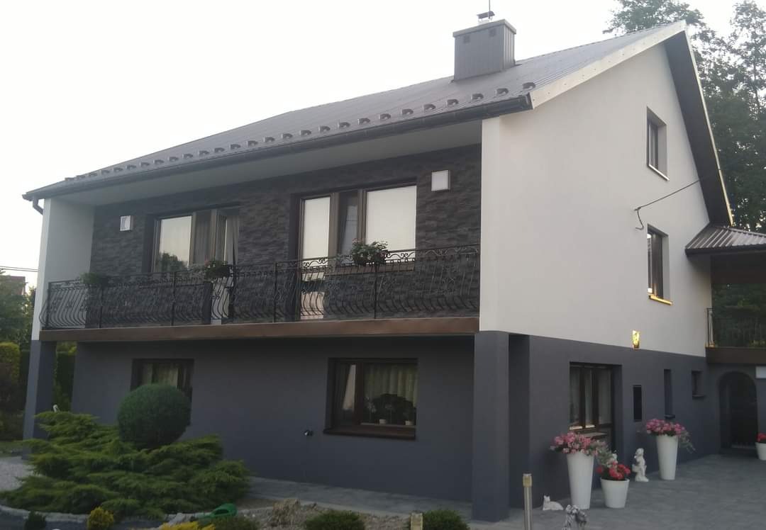 Dwukondygnacyjny dom z elewacją w odcieniach szarości i bieli, z balkonem z kutą balustradą i kwiatami w doniczkach, otoczony zielenią.