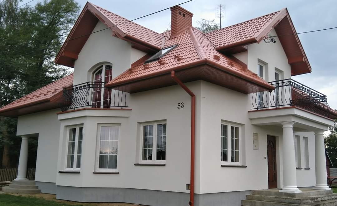Dwupiętrowy dom z białą elewacją, czerwoną dachówką, balkonami z kutymi balustradami i kolumnami przy wejściu, numer 53 na ścianie, widok z zewnątrz.