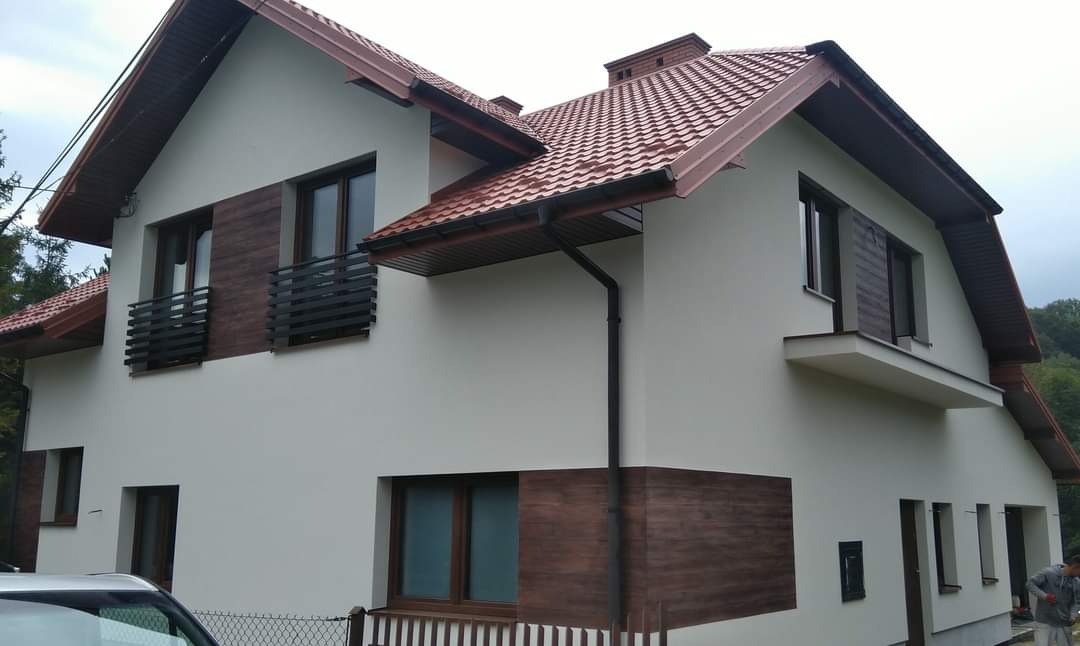 Dwukondygnacyjny dom z jasną elewacją, brązowymi akcentami imitującymi drewno, ciemnobrązową dachówką i czarnymi rynnami, widziany z perspektywy ulicy, z fragmentem samochodu w lewym dolnym rogu.