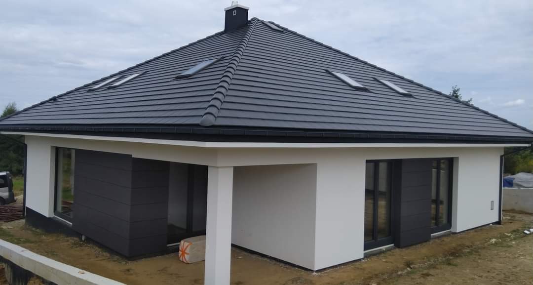 Nowoczesny dom jednorodzinny z grafitową dachówką i fragmentem elewacji wykończonym szarą okładziną, widoczne okna dachowe i komin.