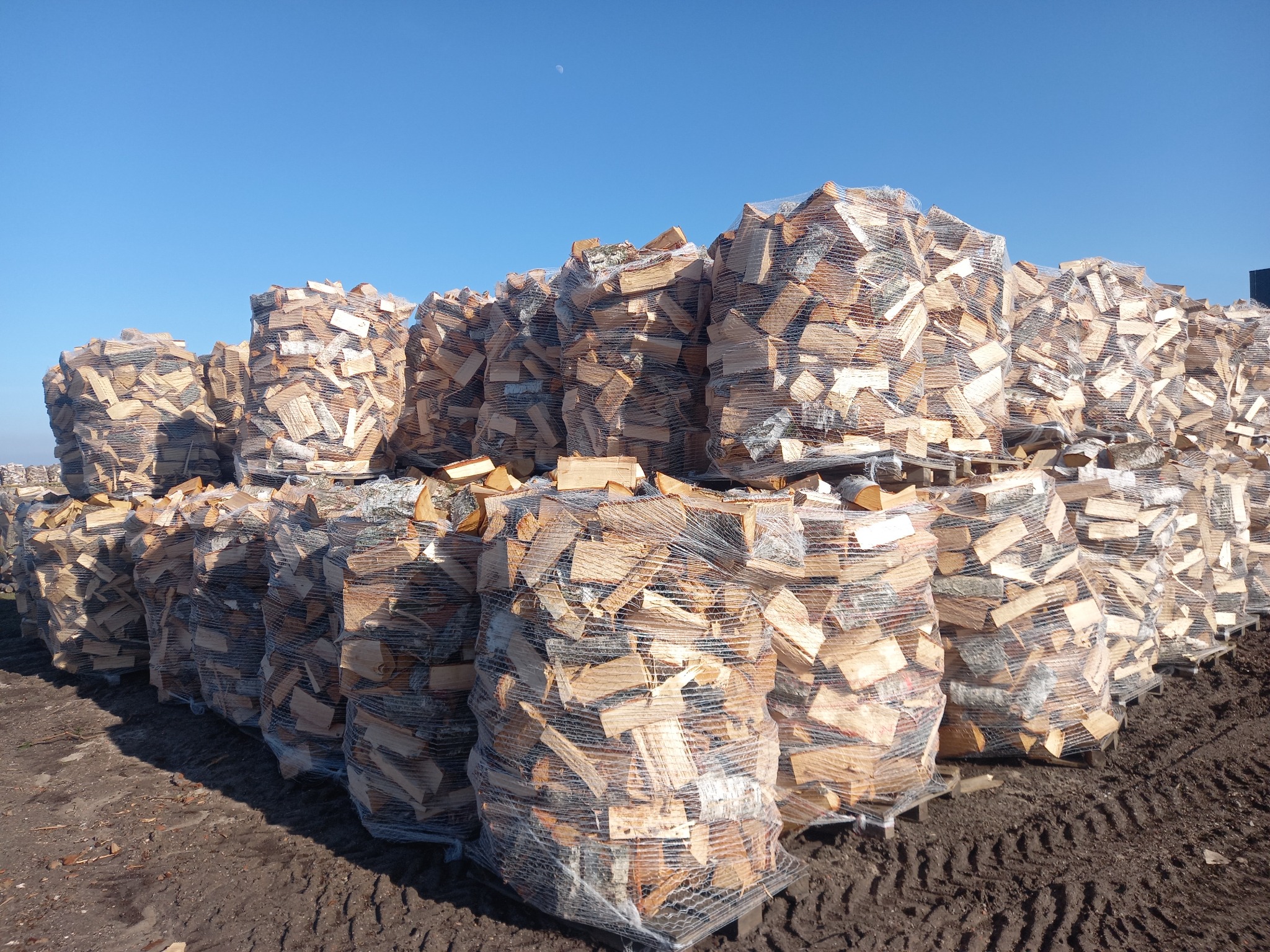 Stosy worków z drewnem kominkowym ułożone na paletach, widoczne ślady opon na ziemi i błękitne niebo z księżycem w tle.