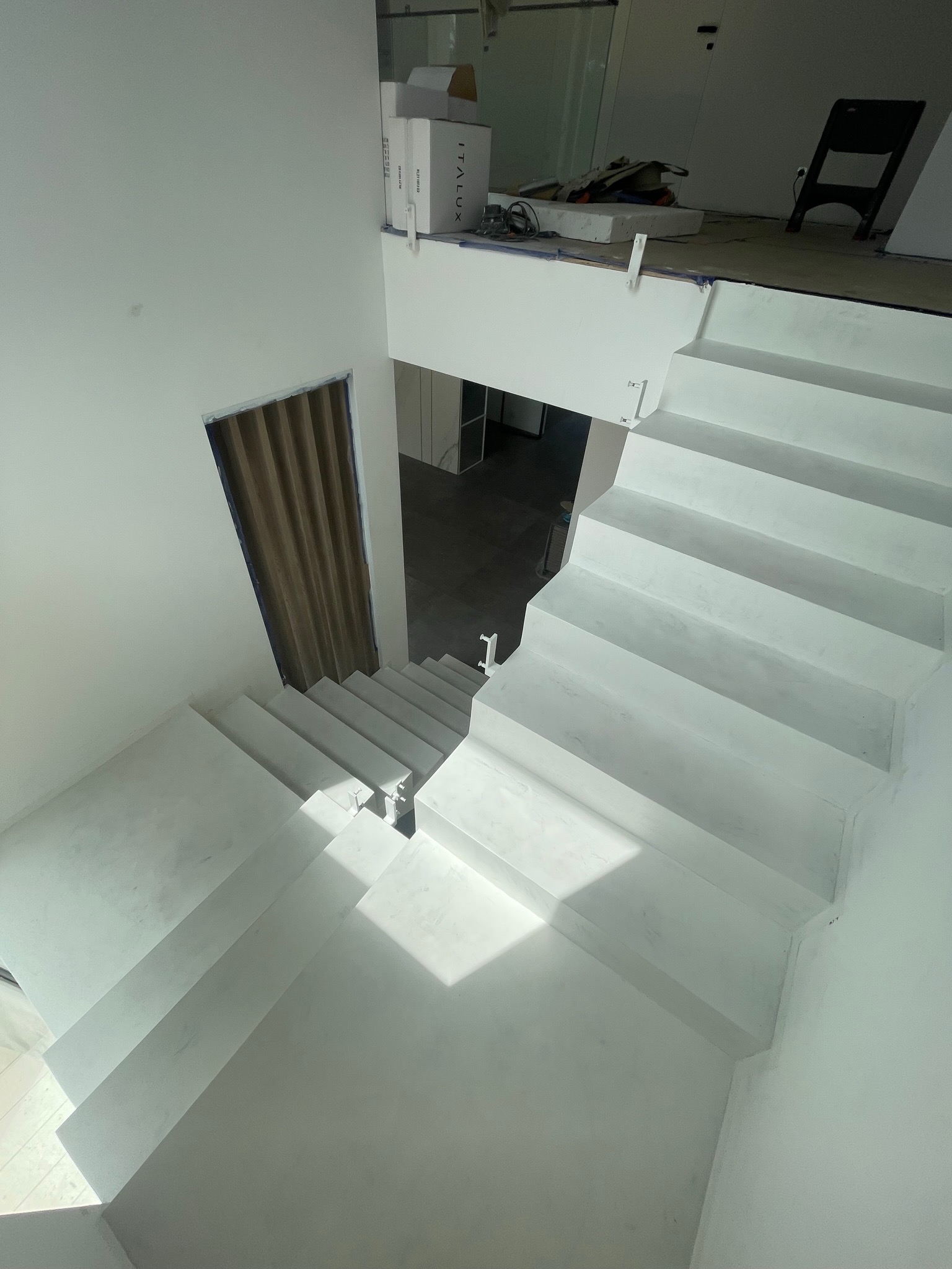 Schody pokryte białym mikrocementem, widok z góry, z widocznym wejściem z drewnianą harmonijką w ścianie, częściowo wykończone pomieszczenie w tle.