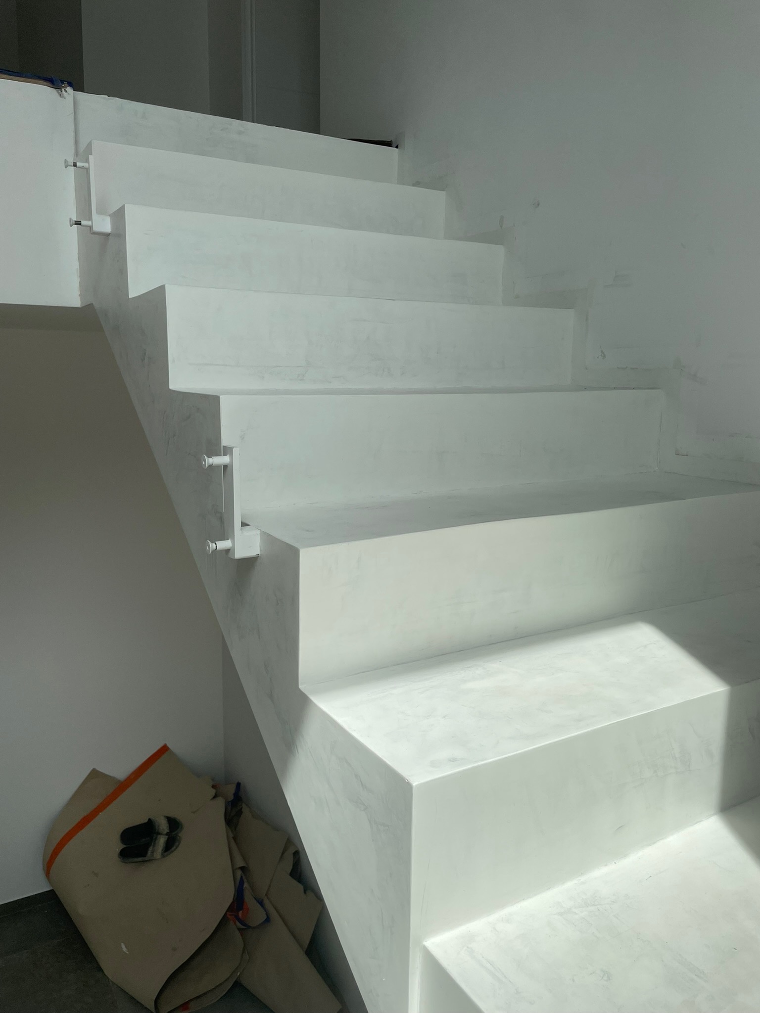 Białe schody pokryte mikrocementem, widoczne ślady szpachlowania i zabezpieczenia krawędzi, z podkładem malarskim, w tle zabezpieczona podłoga.