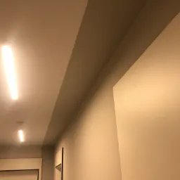 Pomieszczenie z pomalowanymi ścianami w odcieniach beżu, widoczne oświetlenie sufitowe i fragment drzwi.