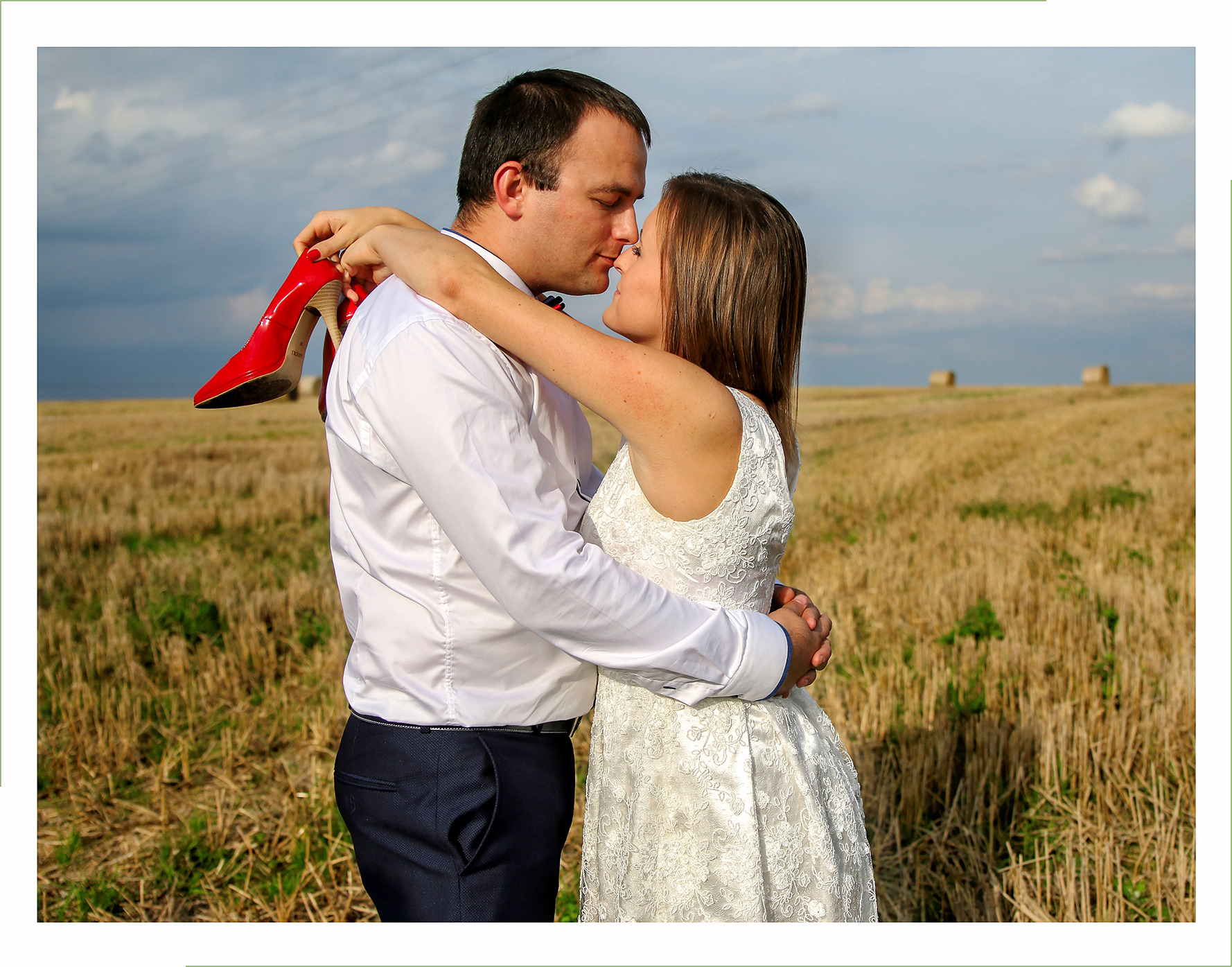 Para w objęciach na polu po żniwach, ona w białej koronkowej sukience, on w białej koszuli i granatowych spodniach, ona trzyma czerwone szpilki na ramieniu, w tle niebo i bele siana.