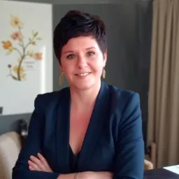 Portret uśmiechniętej kobiety w granatowej marynarce, z założonymi rękami, w tle obraz z motywem roślinnym i zasłona.