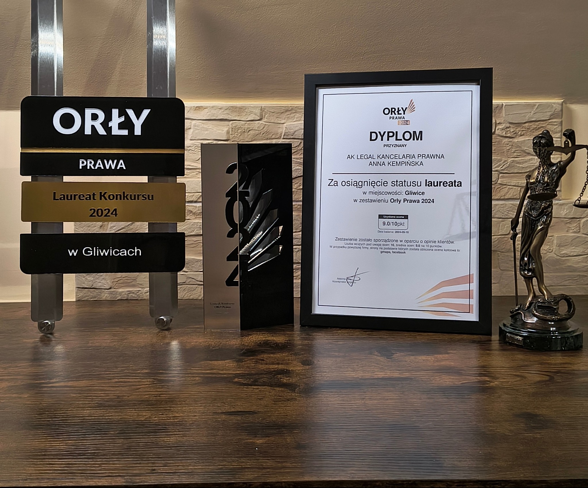 Statuetka i dyplom Orły Prawa 2024 na drewnianym biurku, obok figura Temidy, symbolizujące nagrodę dla kancelarii prawnej.