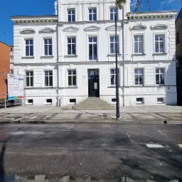 Elewacja odnowionego, trzypiętrowego budynku w stylu klasycystycznym, z licznymi oknami z białymi ramami, czarnymi drzwiami wejściowymi i lampą uliczną przed budynkiem, widziana z perspektywy ulicy...