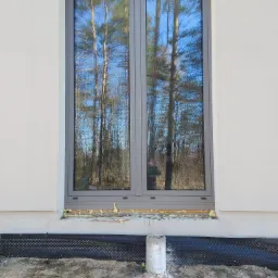 Szare okno z podwójnymi szybami, w których odbija się las i niebo, zamontowane w białej ścianie budynku, z widoczną warstwą izolacyjną i czarną folią fundamentową u dołu.