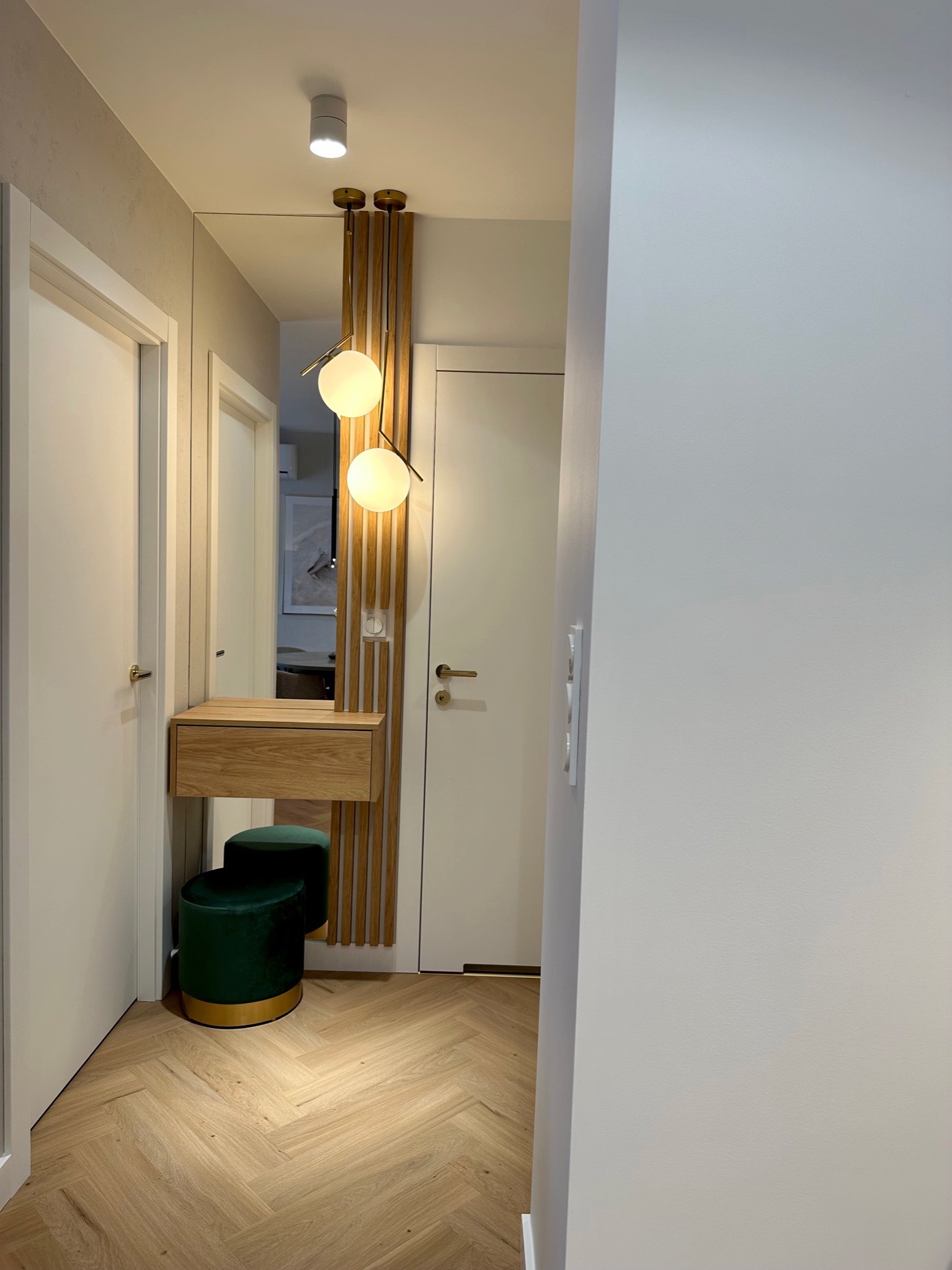 Nowoczesny przedpokój z drewnianą konsolą, okrągłym lustrem, dekoracyjnymi lamelami i zielonym pufem na parkiecie w jodełkę.