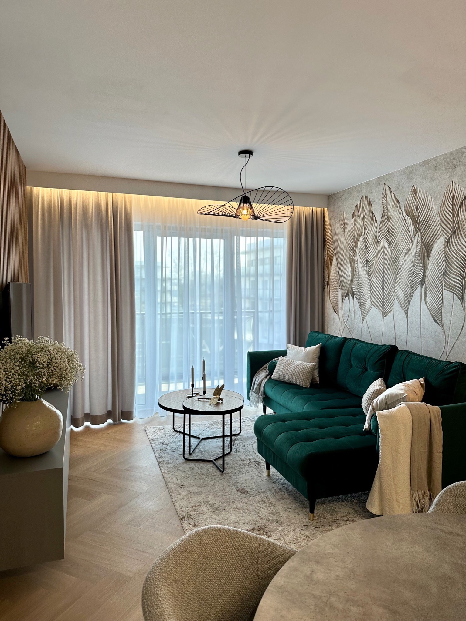 Elegancki salon z zieloną sofą narożną, wzorzystą tapetą w liście, drewnianą boazerią i parkietem w jodełkę, oświetlony lampą z ażurowym kloszem.