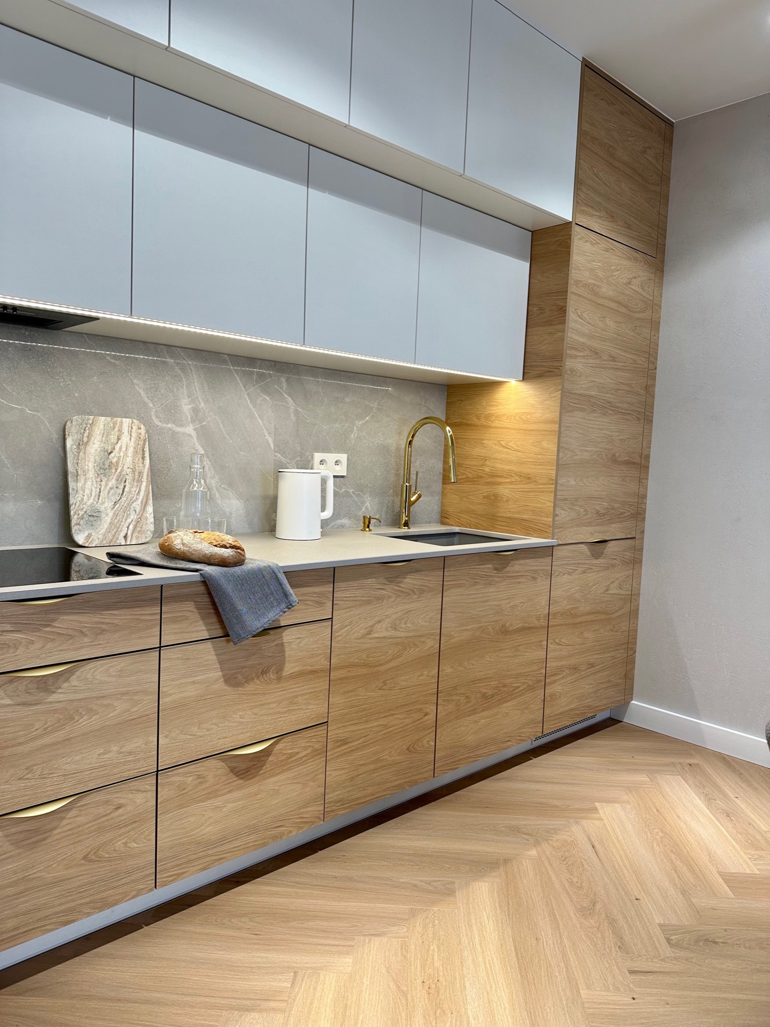 Nowoczesna kuchnia z drewnianymi frontami szafek, złotą armaturą i blatem z szarego marmuru, uzupełniona o dekoracyjne elementy jak chleb, deska do krojenia i szklana karafka.