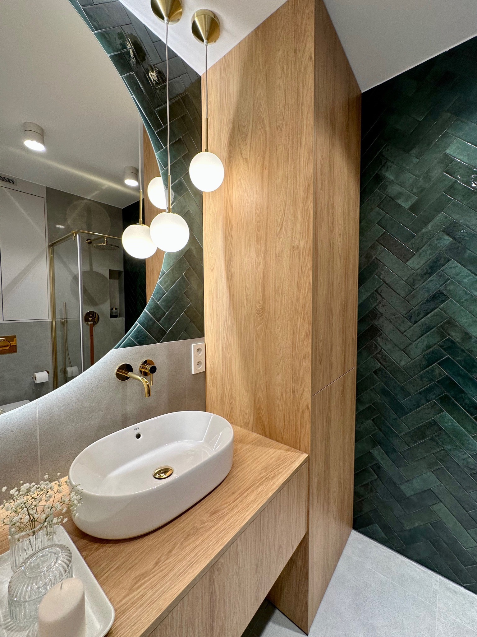Nowoczesna łazienka z umywalką nablatową, baterią w kolorze złotym, oraz ścianą wyłożoną ciemnozielonymi płytkami ułożonymi w jodełkę.