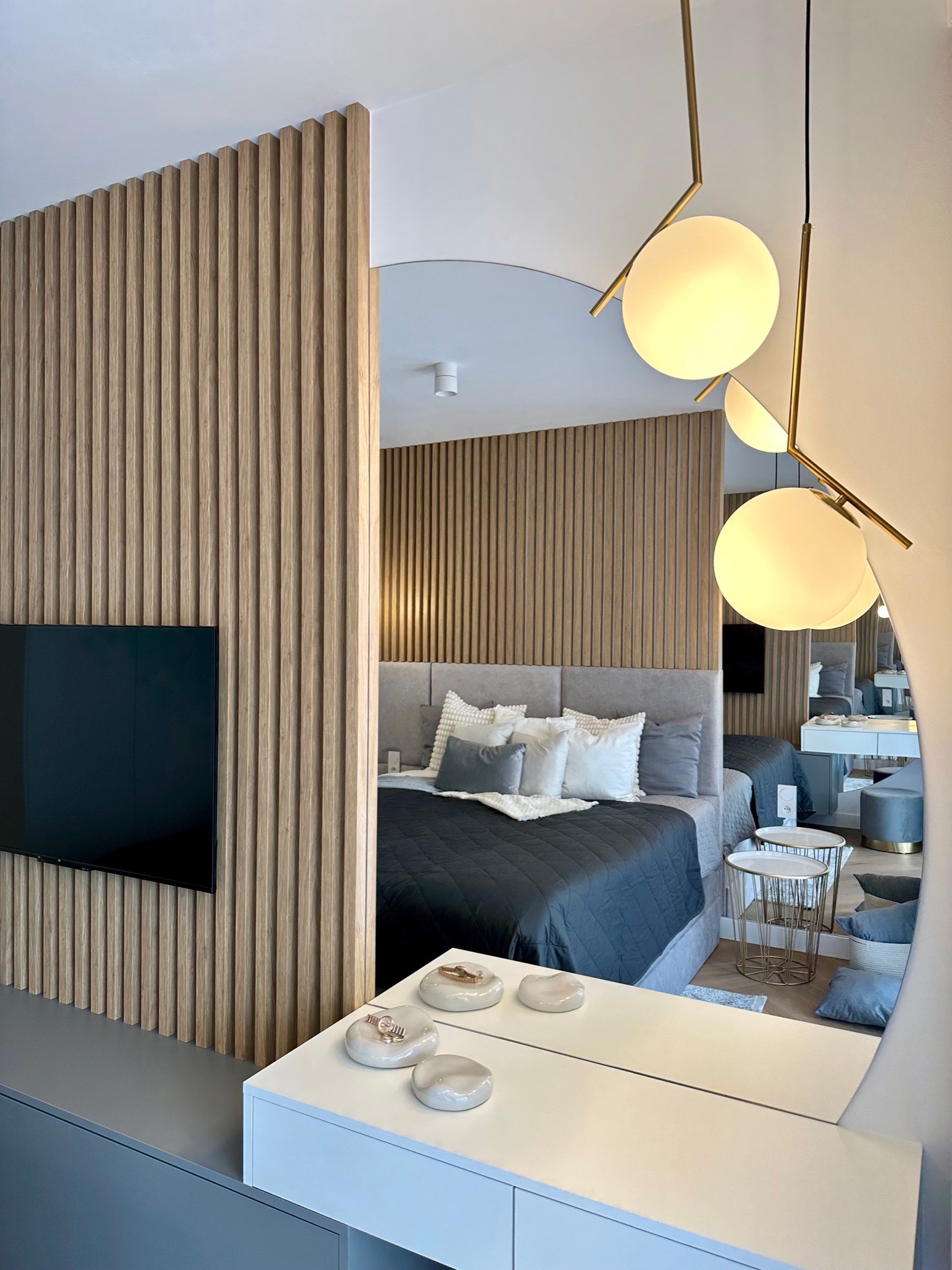Nowoczesna sypialnia z drewnianą ścianką działową, telewizorem i dekoracyjnym oświetleniem, widoczna w lustrze.