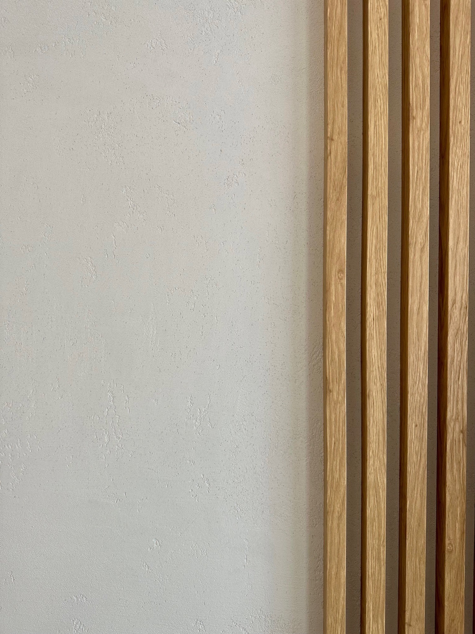 Dekoracyjna ściana z pionowymi drewnianymi lamelami na tle jasnoszarego tynku strukturalnego.