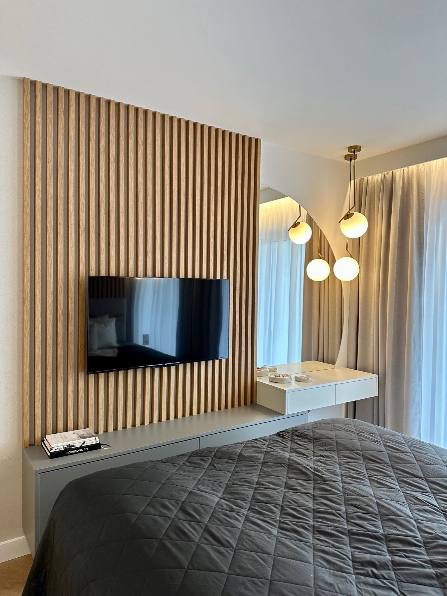 Nowoczesna sypialnia z drewnianą ścianą z lameli, wiszącym telewizorem, szarą szafką RTV, łóżkiem z pikowaną narzutą i toaletką z lustrem oraz designerskimi lampami kulami.